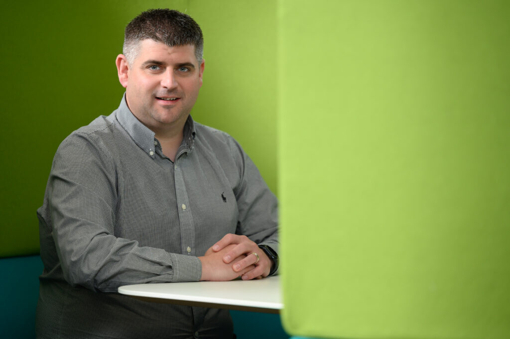 Image of Michael Ryan sitting in front of a green background with his hands folded